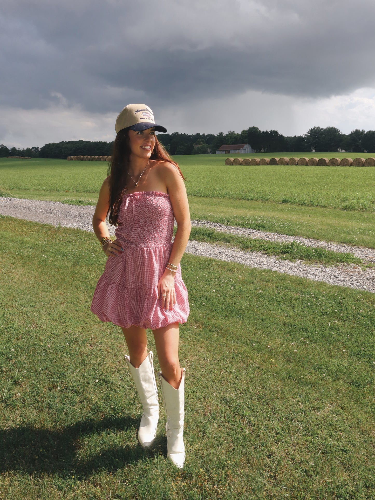 Woman in a pink dress and white boots standing in a field with a cloudy sky.