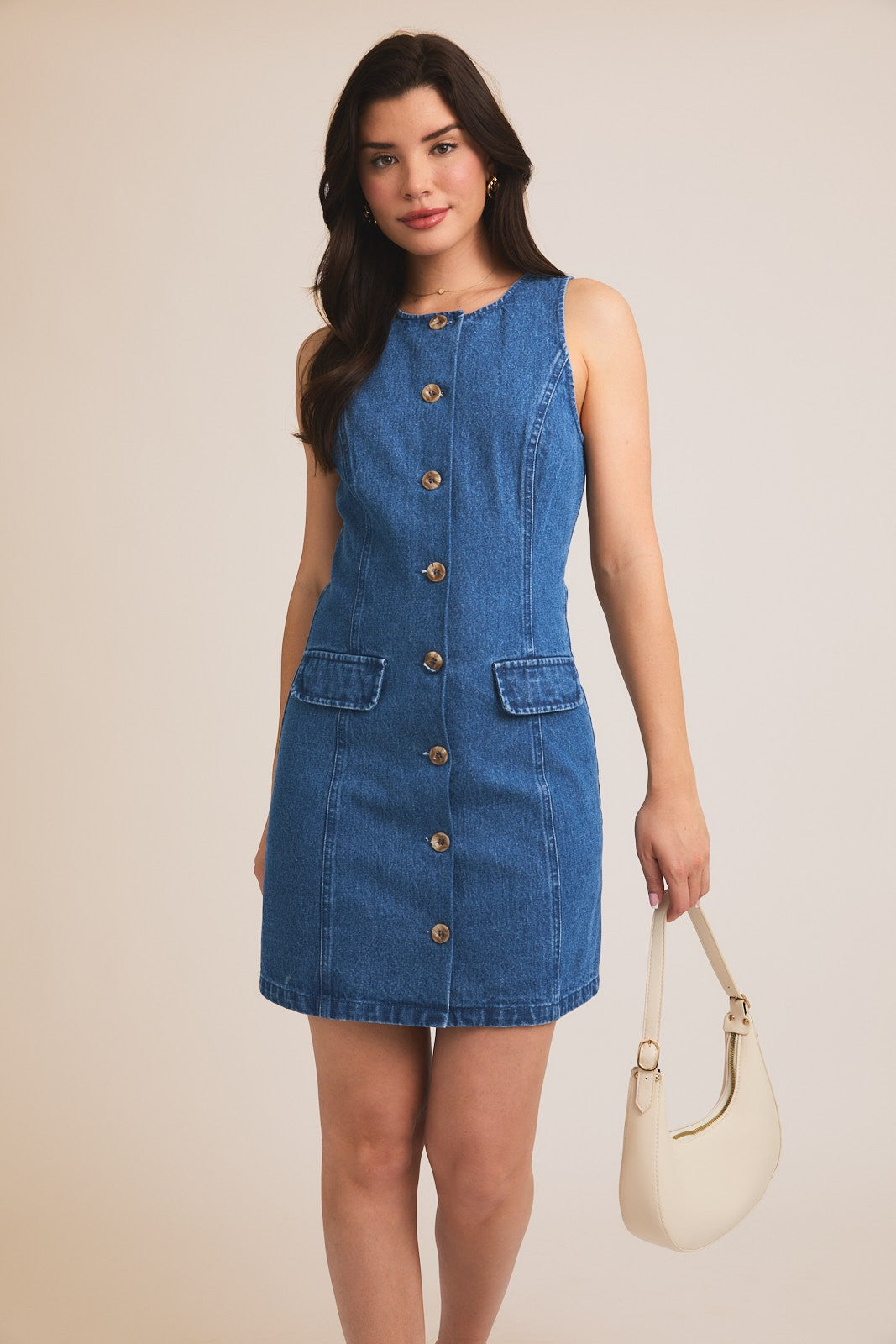 A woman standing against a beige background, wearing a sleeveless blue denim mini dress with button details and pockets.