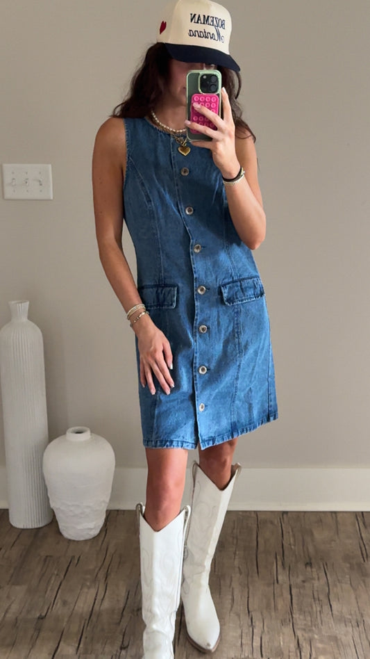 A person wearing a blue denim dress with button details and a round neckline, paired with white boots and a trucker hat.