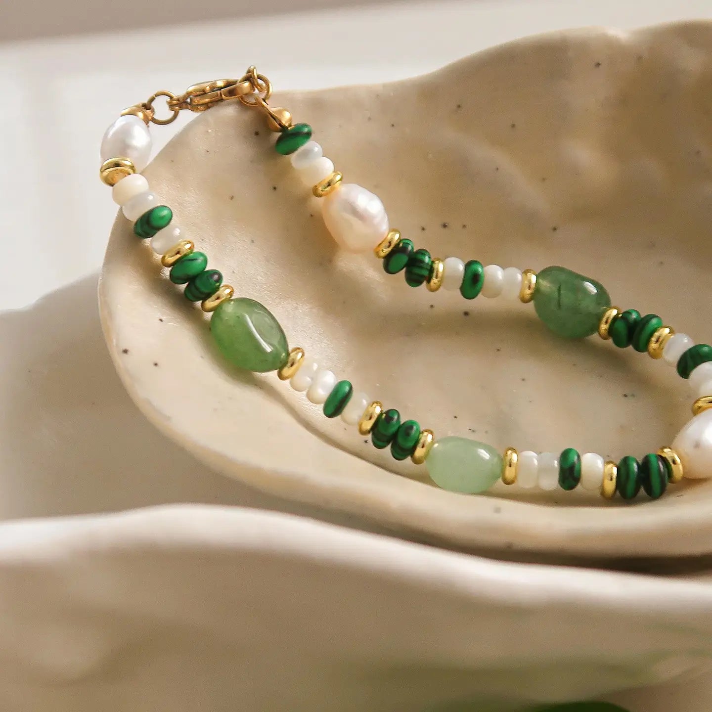 A bracelet with green stone beads and gold-colored accents displayed on a beige ceramic dish.