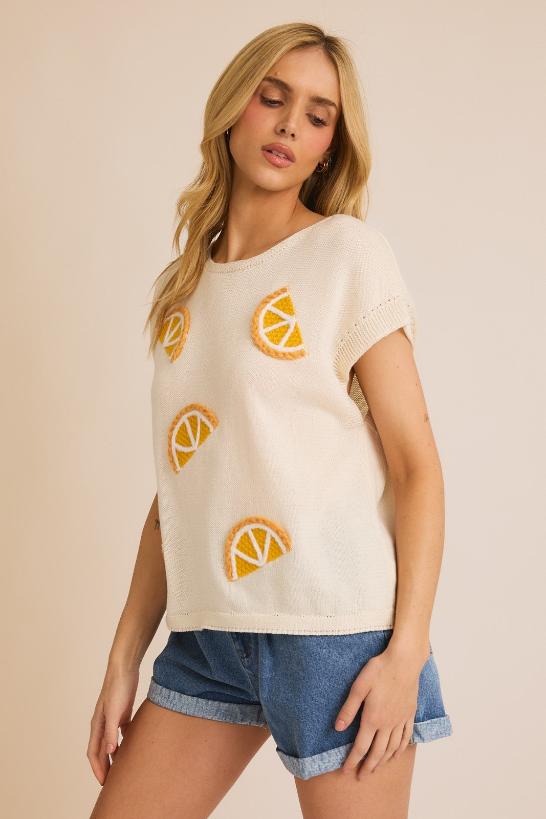 A woman wearing a beige knit top with embroidered lemon designs, paired with blue denim shorts.