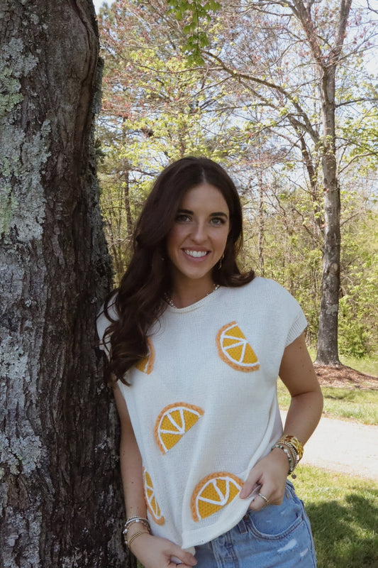 A woman standing outdoors wearing a cream knit top with orange lemon embroidery, paired with blue denim shorts.