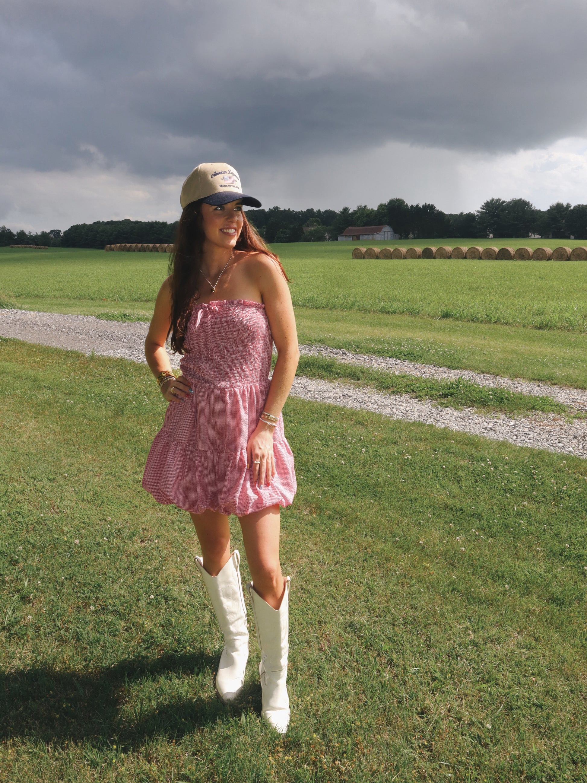 Woman in a pink dress and white boots standing in a field with a cloudy sky.